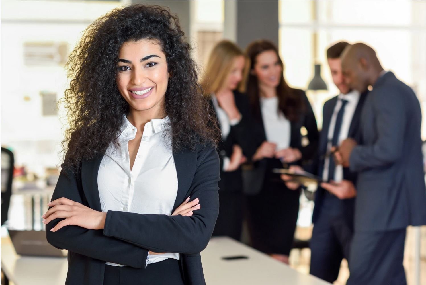 confident-professional-woman-standing-with-arms-crossed-in-modern-office-setting-with-colleagues-blurred-in-background