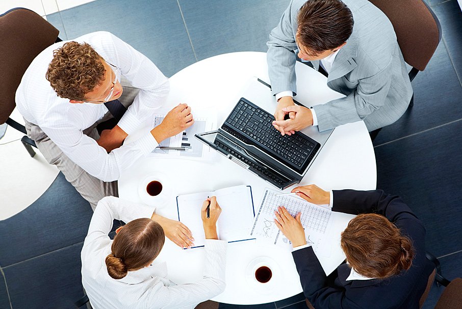 Four business consultants in a collaborative strategy meeting around a white circular table with laptops and notebooks.